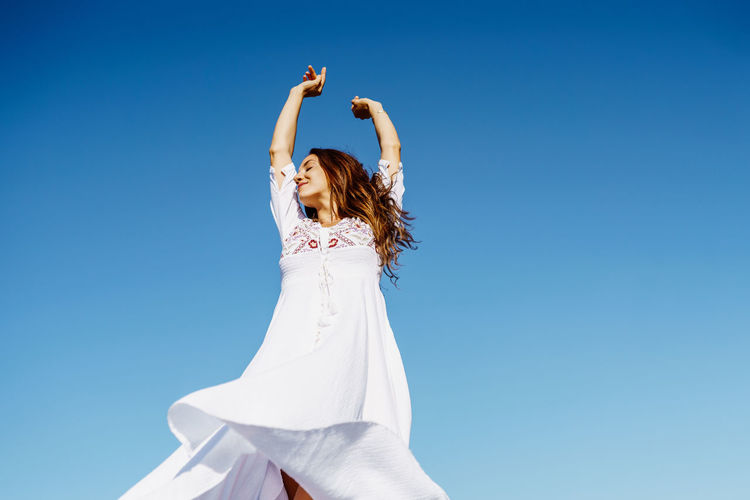 Low angel view of woman with arms raised against clear blue sky