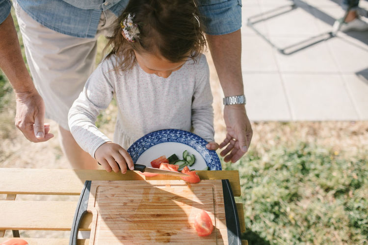Girl putting tomato slices in plate with knife while standing by grandfather at table during garden party