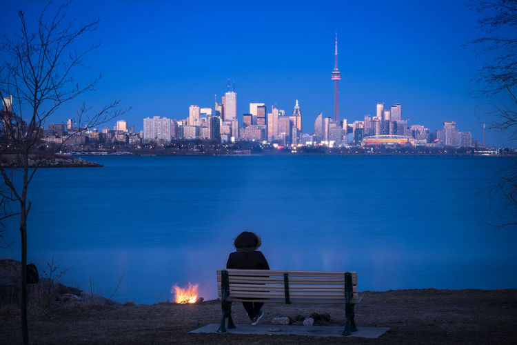 Rear view of woman sitting on bench by river in city at dusk