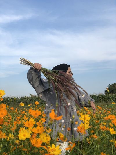 Close-up of young woman holding plants standing at field against sky