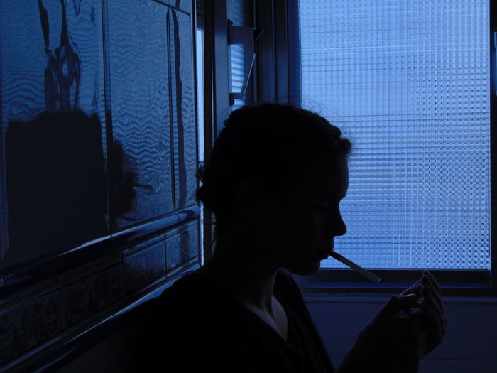 Side view of woman with cigarette while resting by window