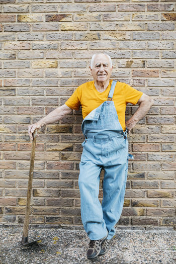 Senior man in denim overall with garden tool looking at camera leaning on a wall