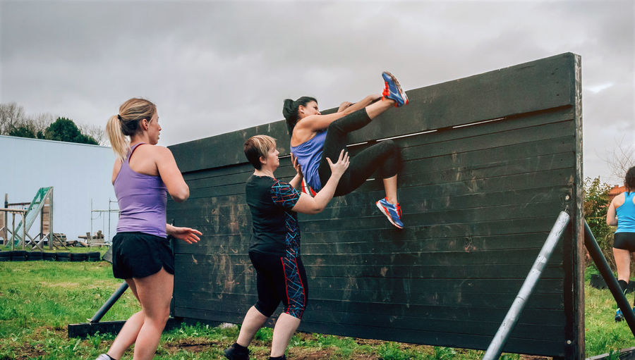 Woman helping friend in climbing wooden wall while exercising outdoors