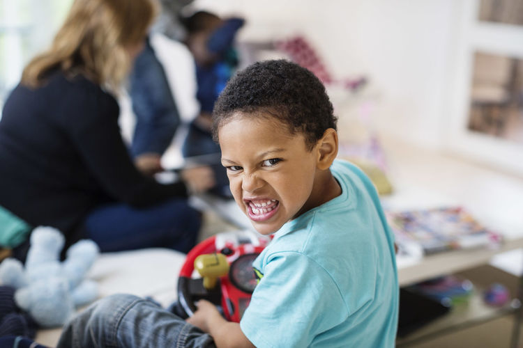 Portrait of boy making face while playing at home with mother sitting in background
