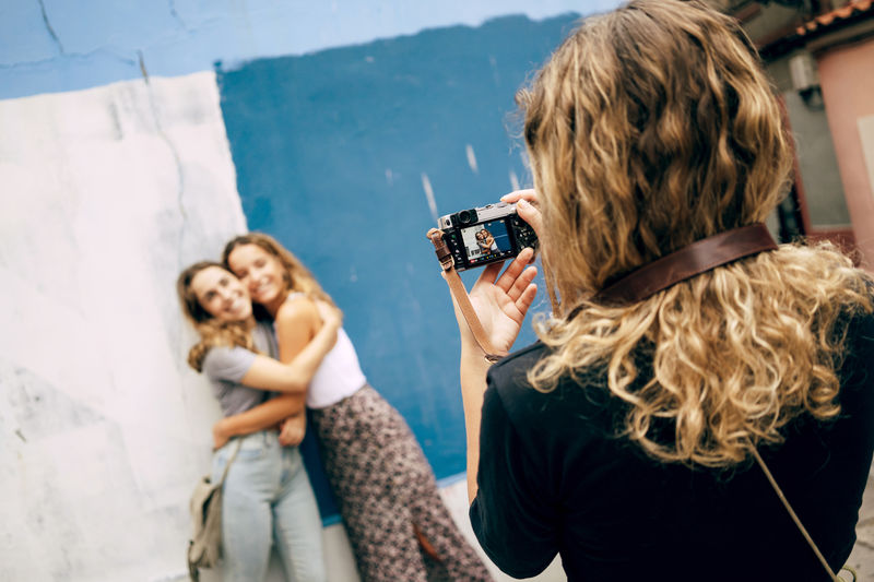 Charming friends hugging posing against wall while woman taking picture on camera