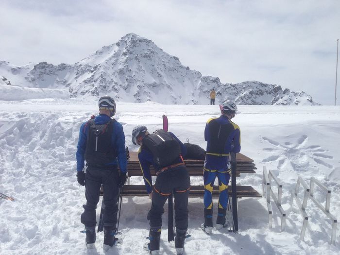 Rear view of skiers standing by wooden bench on snow covered field against mountain