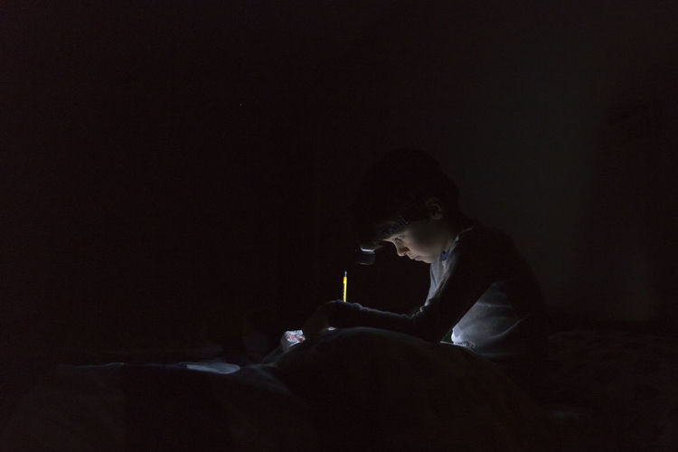 Side view of boy with illuminated headlamp writing homework while sitting on bed in darkroom at home