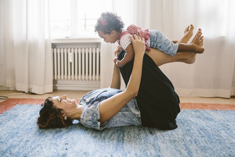 Playful mother lifting daughter while lying on carpet at home