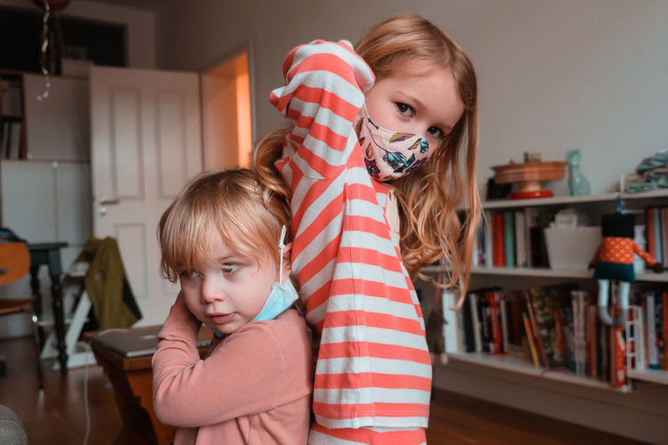 Sisters wearing face masks while standing back to back at home