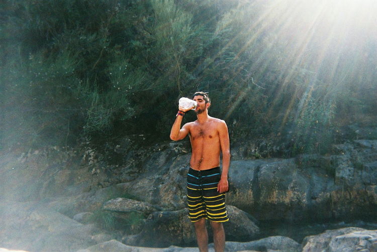 Young man drinking water from bottle by rocks