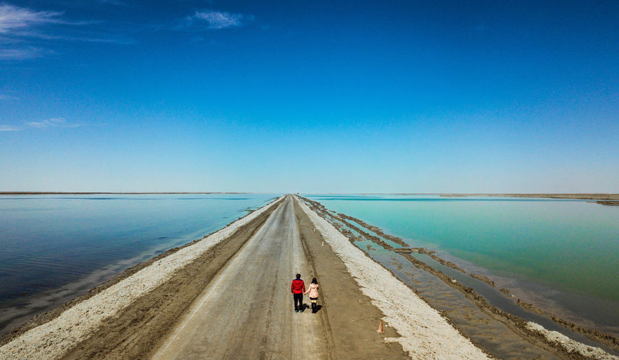 Rear view of man and woman walking on road amidst sea against blue sky