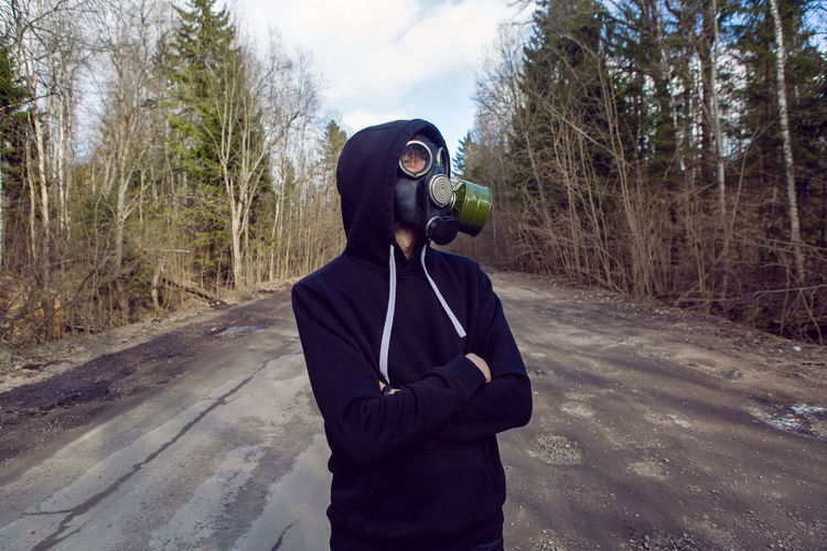 Guy stands in a post apocalyptic forest during the day wearing a gas mask during a coronavirus