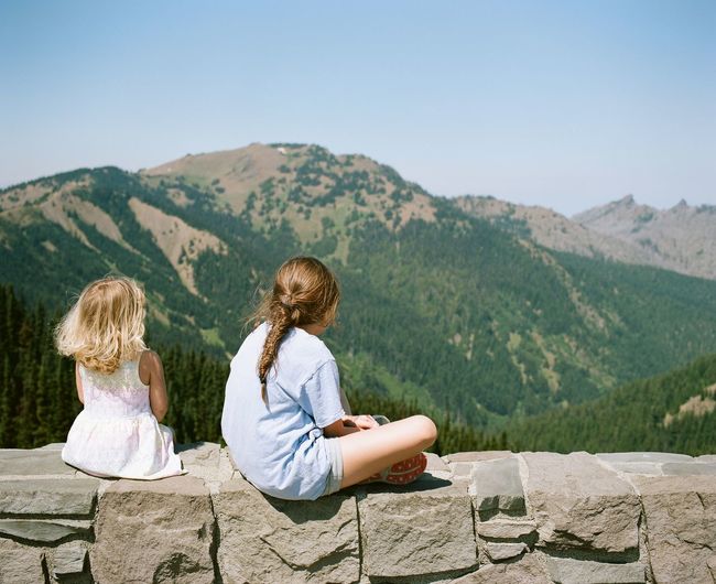 Rear view of girls sitting on stone wall against mountain