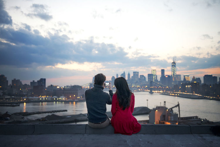 Rear view of couple clicking photograph of illuminated cityscape at dusk