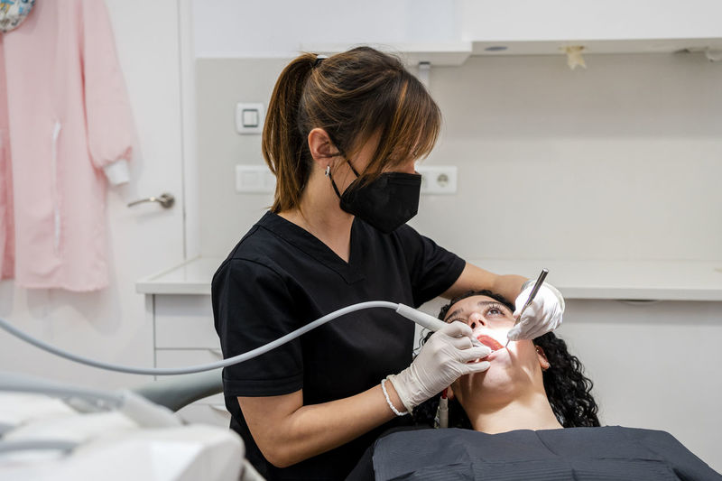 Female dentist wearing protective face mask doing dental suction of patient in clinic