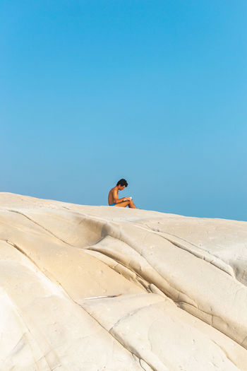 Shirtless man sitting on sand dune against clear blue sky