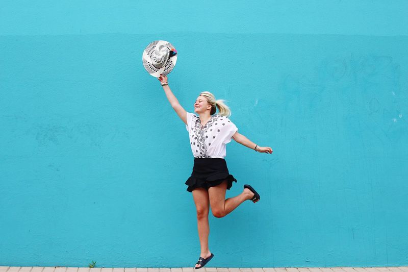 Full length of young woman holding hat while jumping against blue wall