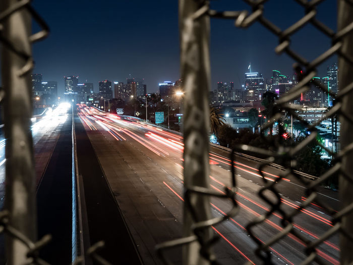 High angle view of light trails on roads in city seen through broken chainlink fence at night