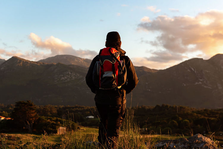 Back view of unrecognizable male traveler with backpack standing on hill and admiring scenery of mountain ridge under sunset sky