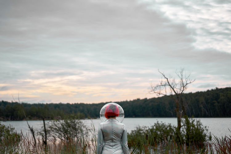 Back view of unrecognizable futuristic young red haired female in silver space suit and glass helmet walking to flooding river