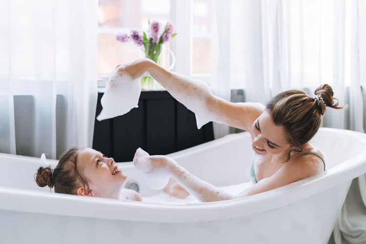 Young mother woman with little girl daughter in pajamas having fun in the bath with foam at home