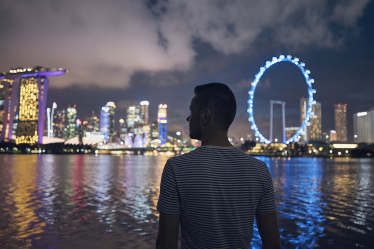 Man standing on waterfront of bay and looking at city at night. urban skyline with bokeh of lights.
