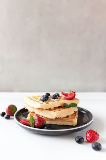 Stack of waffles on plate on white table, blueberry, chopped strawberry, mint leaf, gray background