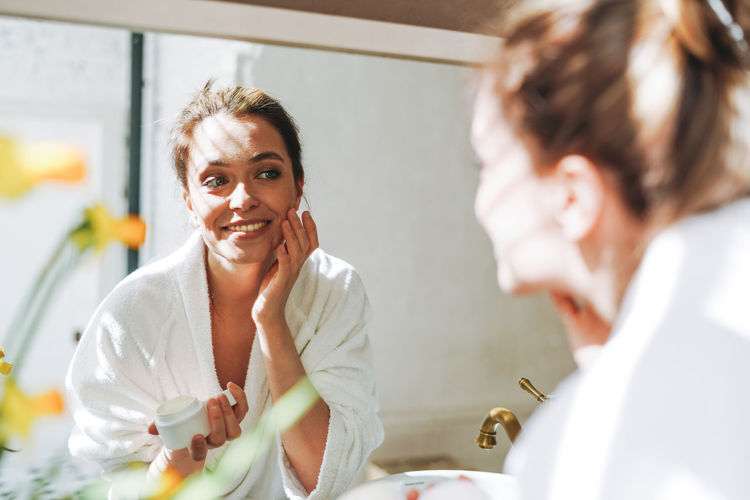 Young woman in white bathrobe put cream on clean fresh skin face near mirror in bathroom at home