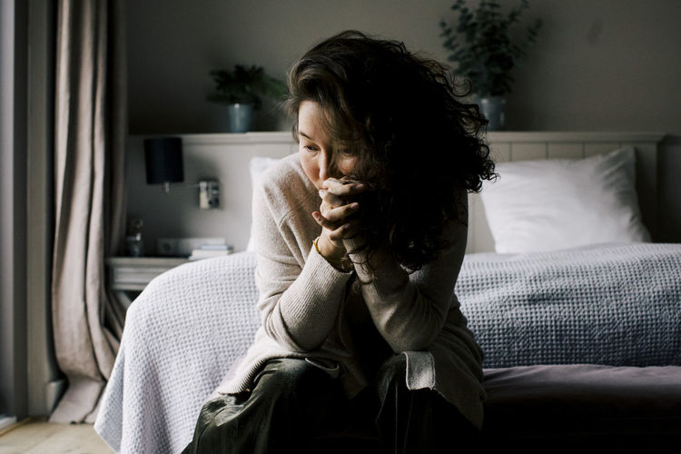 Depressed woman sitting with hands clasped contemplating in bedroom at home