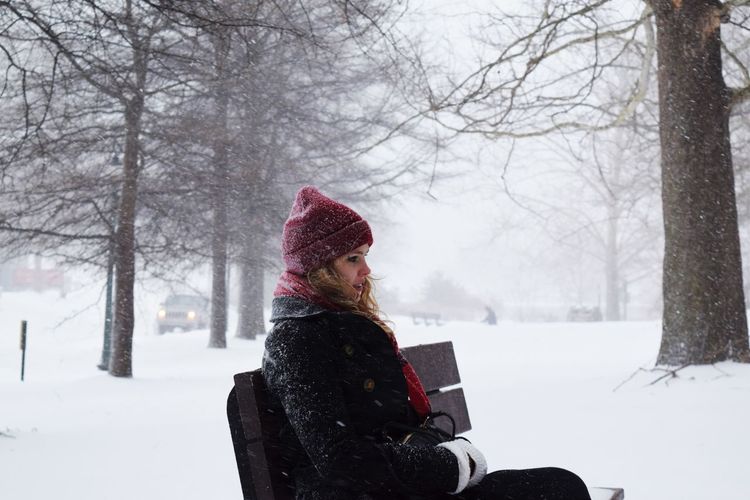 Side view of young woman sitting on bench at park during snowfall
