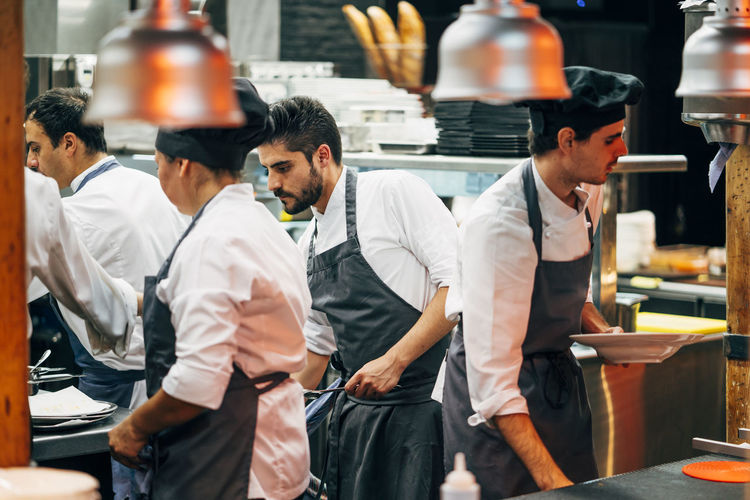 Men in hats and aprons working in team and serving dishes with delicious food in kitchen