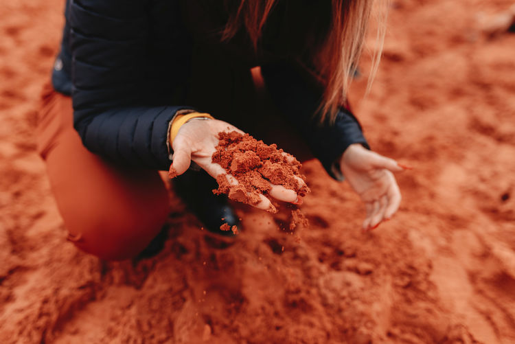 From above of crop unrecognizable female sitting on haunches in warm clothes touching smooth red sand in desert