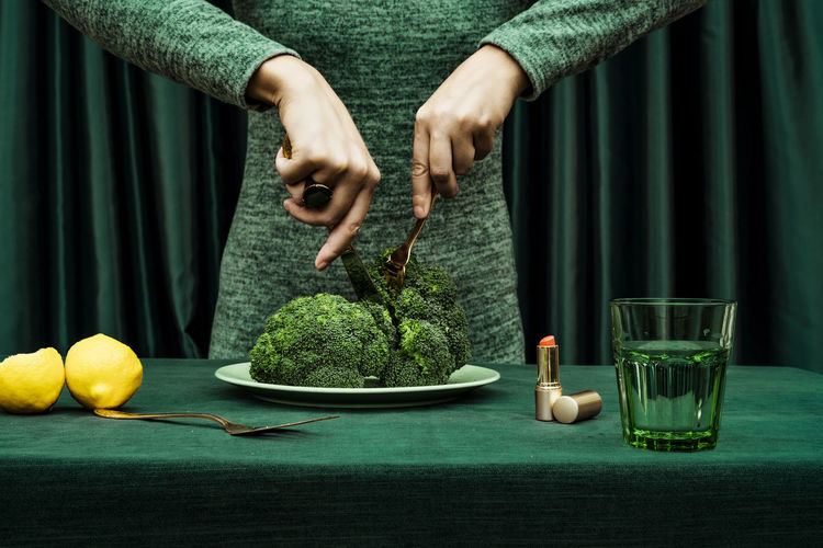 Midsection of woman cutting broccoli with knife and fork while standing against green curtain