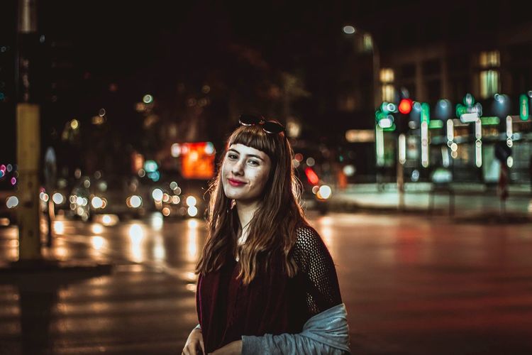 Portrait of smiling teenage girl standing on street in illuminated city at night