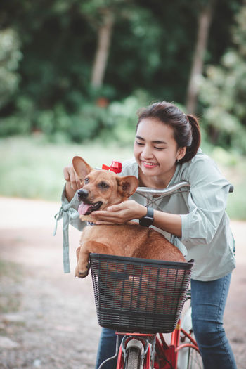 Cheerful girl with dog in basket of bicycle standing outdoors