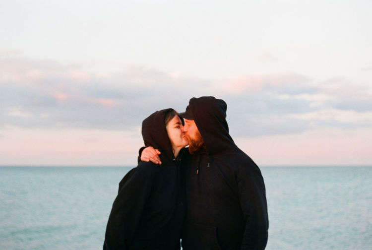 Young kissing couple standing against sea against sky during sunset