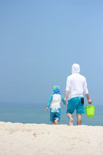 Rear view of father and son walking on beach against clear blue sky