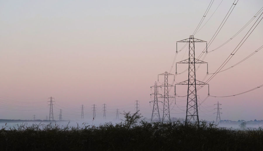 Low angle view of electricity pylons on field against sky during sunrise