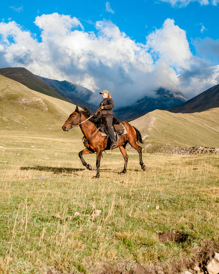 Side view of a cowgirl shepherd riding on a horse on a mountain valley against sky with clouds