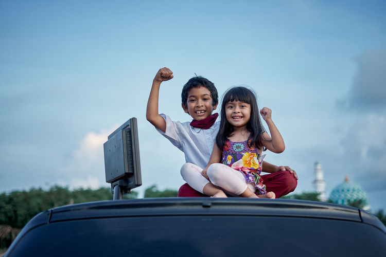 Portrait of smiling siblings sitting on car against sky