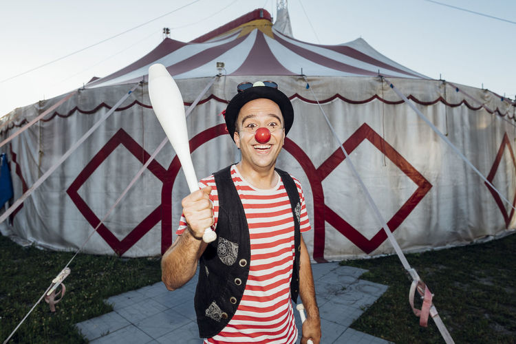 Smiling male clown holding juggling pin while standing in front of circus tent