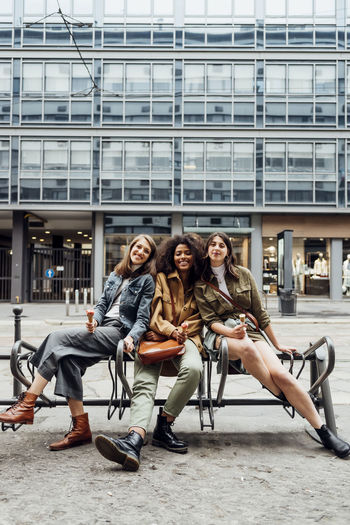 Smiling female friends sitting together on bench in city