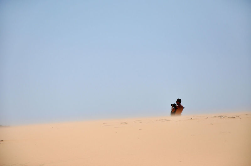 Young man photographing while standing in desert against clear sky