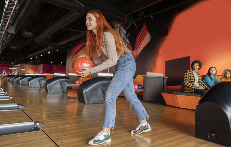 Young woman throwing ball with friends sitting in background at bowling alley