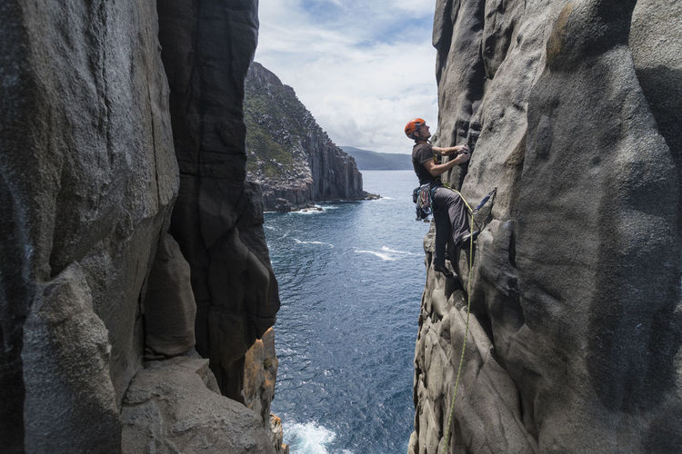 Daring man rockclimbs the exposed edge of a dolerite rock ridge, with the ocean seen in the background in cape raoul, tasmania, australia.