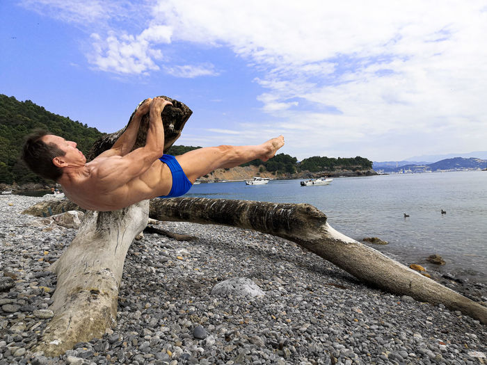 Full length of shirtless man exercising on driftwood at sea