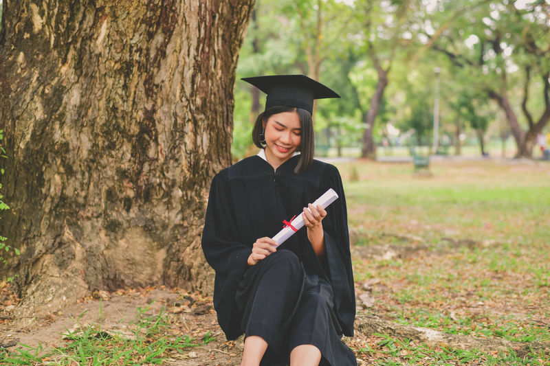 Young woman in graduation gown sitting on field at park