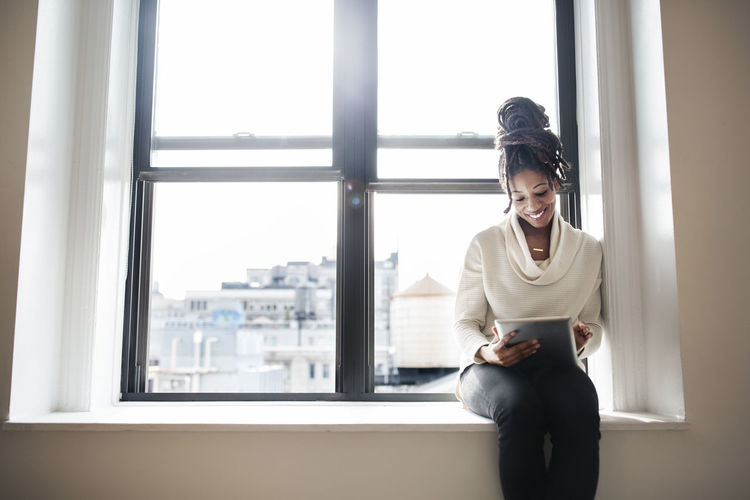 YOUNG WOMAN USING PHONE WHILE SITTING ON WINDOW AT HOME