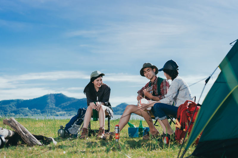 Friends looking at young man playing guitar while sitting on chairs against sky