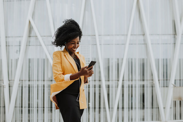Smiling female entrepreneur using smart phone listening to music through earphones standing against building